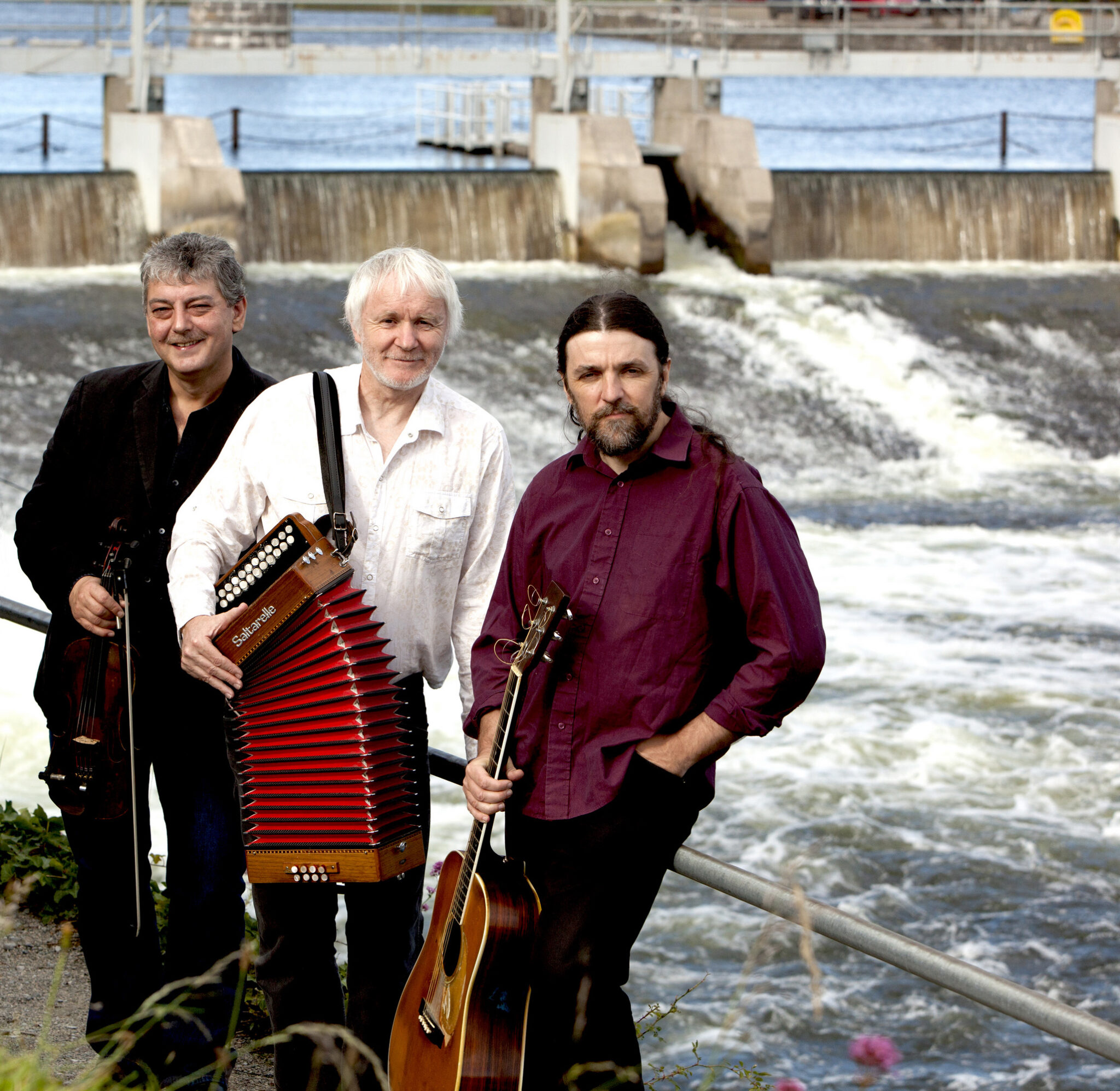 three men sit in front of a dam or waterfall with instruments